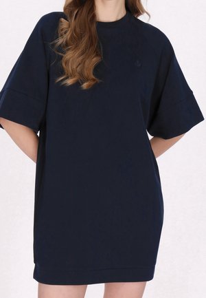 Femme portant une robe ample bleu marine foncé jusqu'aux genoux avec des manches courtes larges et de longs cheveux ondulés sur l'épaule gauche.