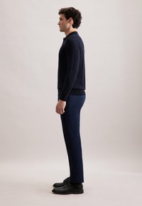 Granatowy sweter z dzianiny, dopasowane granatowe spodnie i czarne skórzane buty. Model stoi w profilu na neutralnym tle.