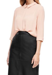 Femme portant un chemisier à manches longues rose clair rentré dans une jupe en cuir noire taille haute avec des coutures visibles.