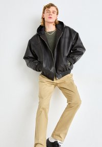 Svart lær zip-up jakke med hette, ribbede mansjetter og lommer, båret over en grønn genser og beige bukser. Modell som står mot en hvit bakgrunn.