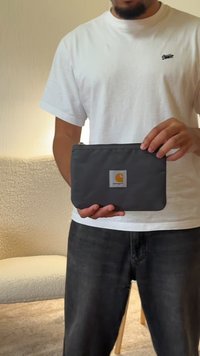 Pochette en tissu gris avec une fermeture à glissière, arborant une étiquette carrée du logo Carhartt en orange et gris. Forme rectangulaire, texture lisse.