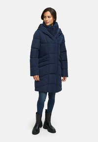 Marineblauwe gewatteerde jas met een brede capuchon, ritssluiting en zijzakken. Heeft een doorgestikte textuur en wordt gecombineerd met donkere skinny jeans en zwarte laarzen.