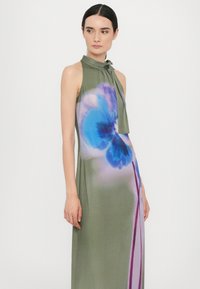 Lange, mouwloze jurk in olijfgroen met een hoge hals en strikdetail. Heeft een bloemenprint in blauwe en paarse tinten aan de voorkant.