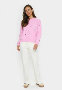 Roze sweatshirt met rode hartpatronen, ronde hals, geribbelde mouwen en onderkant, gecombineerd met witte wijde broek en beige sneakers.