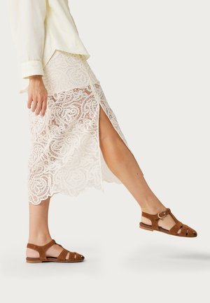 Mujer con falda midi de encaje blanca con abertura lateral y sandalias planas marrones con tira en T, levantando una pierna hacia adelante sobre un fondo neutro.