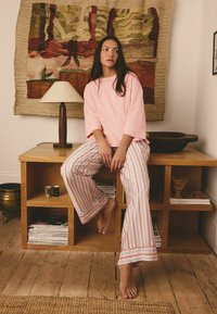 Kobieta w różowej bluzce i paskowanych piżamowych spodniach siedzi boso na drewnianej półce w kształcie kostki, za nią lampa i wiszący na ścianie wieszak.