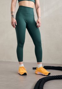 Zielone legginsy o wysokim stanie z gładką teksturą, w połączeniu z jasnobrzoskwiniowymi butami sportowymi z czarnymi akcentami, na neutralnym tle.