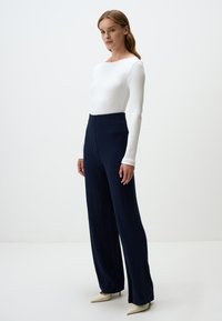 Maglietta bianca a coste a maniche lunghe abbinata a pantaloni larghi a pieghe blu navy e décolleté beige a punta, su uno sfondo bianco uniforme.