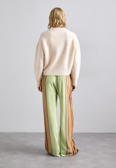 Beige, pörröinen neule, jossa on rento mitoitus. Vaaleanvihreät, leveälahkeiset housut, joissa on pystysuorat raidat oranssilla, ruskealla ja tummanvihreillä yksityiskohdilla.