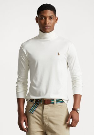Witte coltrui met lange mouwen, voorzien van een klein logo op de borst. GeCombineerd met beige broek en een meerkleurige gestreepte riem.
