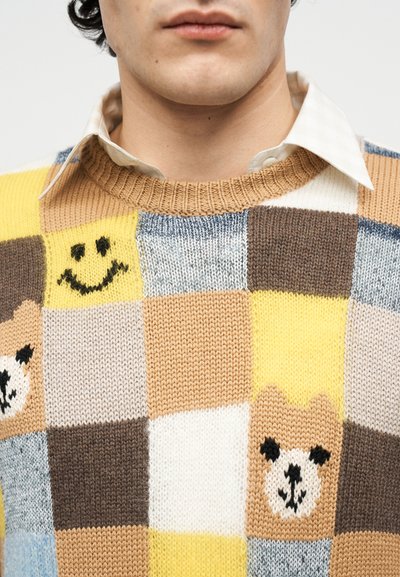 Άνδρας που φοράει πλεκτό πουλόβερ παζλ με κίτρινα, καφέ και μπεζ τετράγωνα και σχέδια από χαμογελαστά πρόσωπα και κεφάλια αρκούδας, φορεμένο πάνω από λευκό πουκάμισο με γιακά.