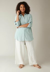 Hellblaues Button-Up-Hemd mit hochgerollten langen Ärmeln, kombiniert mit weißen Leinen-Hose in weitem Schnitt und braunen Sandalen mit Riemendetail.