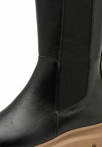 Botte en cuir noir avec des panneaux latéraux élastiques, une texture lisse et une semelle en caoutchouc beige avec des détails striés pour une bonne traction.