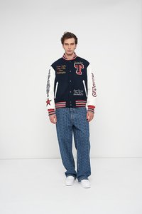 Temno modra varsity jakna z belimi rokavi, rdečimi detajli in izvezenim besedilom. Par svetlo modrih kavbojk z vzorcem in bele superge.