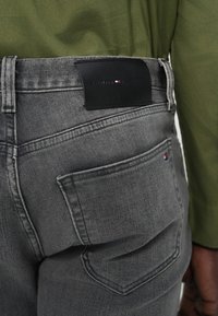 Γκρι τζιν από denim με μαύρη δερμάτινη ετικέτα στη μέση και πίσω τσέπη με μικρό πολύχρωμο λογότυπο. Η υφή είναι λεία με ορατή ραφή.