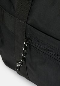 Mochila de tecido preto com um bolso com fecho, material texturizado, alças trançadas e um cordão de fecho com padrão contrastante.