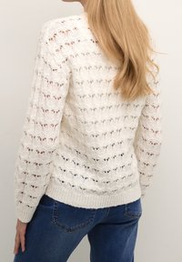 Suéter blanco de punto con un patrón de calado, mangas largas y un dobladillo acanalado. Combinado con jeans azules, mostrando un ajuste relajado.