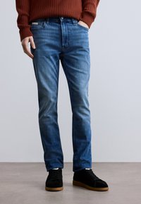 Kék farmer, karcsúsított szabás, kopott hatású mosás és kontrasztos varrások. Fekete texturált tornacipővel és bordázott, rozsaszín pulóverrel párosítva.