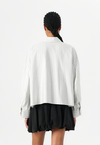 Chemise oversize rayée gris clair avec des épaules tombantes, des poignets boutonnés et un ourlet court, assortie d'une jupe noire à plusieurs couches.