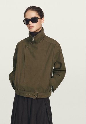 Giacca verde oliva con colletto alto, chiusura a scatto e vita cinturata, abbinata a un maglione nero a collo alto e occhiali da sole neri oversize.