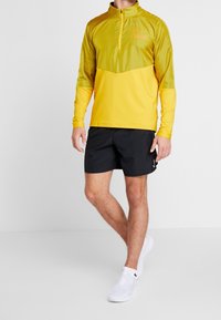 Camiseta atlética de manga larga amarilla con diseño de media cremallera, que presenta paneles texturizados en los hombros; combinada con pantalones cortos negros y zapatillas blancas.