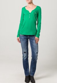 Top verde de manga larga con escote en V y tapeta de botones, combinado con jeans azules desgastados y botas negras de tobillo. Tejido suave y diseño ajustado.