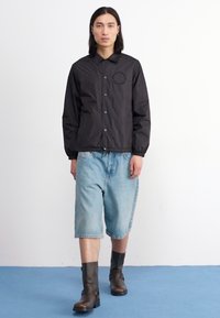 Black, lightweight jacket with a snap-front closure and round logo detail, paired with loose-fitting light blue denim shorts and chunky brown boots.