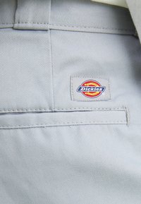 Sivkasta tkanina z vidno žepek in prišitimi oznako Dickies v rdeči, rumeni in modri barvi. Tekstura je gladka in trpežna.