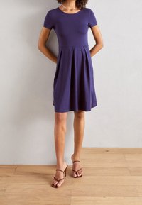 Abito navy a maniche corte con corpino aderente e gonna svasata. Realizzato in tessuto morbido, presenta un collo rotondo e un design plissè. Sandali con tacco marroni.