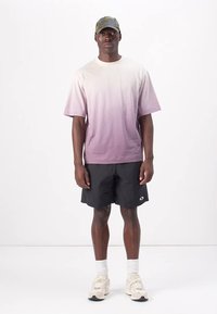 Kurzärmliges T-Shirt mit Farbverlauf von Weiß zu Pink, kombiniert mit schwarzen Shorts. Das Outfit wird mit einer Camouflage-Mütze und hellen Sneakers abgerundet.