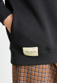 Sweatshirt negru cu o textură ribforată, buzunar tip cangur și un etichetă crem cu inscripția "STUDIO EDITION" la margine. Pantaloni căptușiți maro în carouri dedesubt.