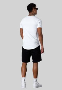 Weißes Baumwoll-T-Shirt mit kurzen Ärmeln, abgerundetem Saum, getragen mit schwarzen Shorts. Weiße Sneakers und gemustertes Socken. Schlichtes Design, keine Grafiken.