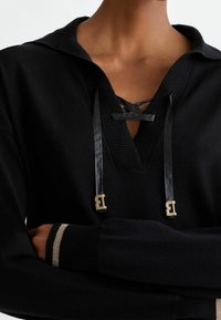 Czarny pulower z kapturem, z sznurowanym dekoltem i błyszczącymi czarnymi sznureczkami. Złote akcenty oraz ściągacze z metalicznym paskiem.