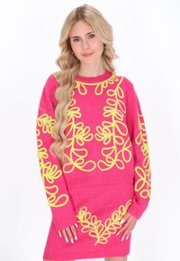 Jasnoróżowy sweter z dużym żółtym haftem w kwiaty, z prążkowanym dekoltem i mankietami. Miękka faktura. Modelka pozuje z rękami na biodrach.