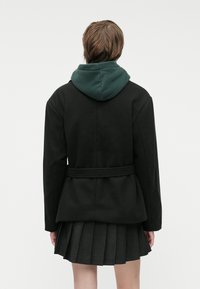 Black coat with a green hood, featuring a belted waist, structured design, and pleated skirt below. Smooth fabric and tailored fit.