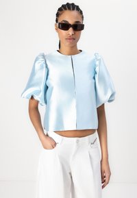 Lyseblå satinskjorte med cropped design og puffermer, med glidelås foran og en glatt, skinnende tekstur. Kombinert med hvite bukser.