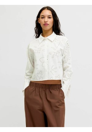 Kvinne iført en hvit langermet øyeskapsknappet skjorte og brun høytlivsbukse med hendene i lommene, står foran en ensfarget bakgrunn.