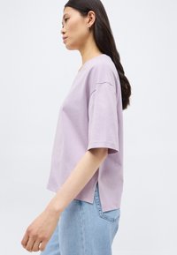 Νεαρή γυναίκα με μακριά σκουρόχρωμα μαλλιά, ντυμένη με μια λιλά oversized μπλούζα και ανοιχτόχρωμο τζιν, στέκεται προφίλ σε λευκό φόντο.