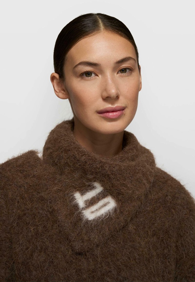 Brązowy sweter z mieszanki wełny z szerokim kołnierzem, zdobiony kremowym wzorem. Materiał o fakturze puchatej. Starannie oprawiona twarz w neutralnych tonach.