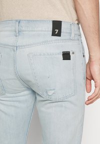 Pantalones vaqueros de mezclilla azul claro con un acabado desgastado, que cuentan con bolsillos traseros, una etiqueta de marca negra y un detalle desgastado cerca del bolsillo derecho.