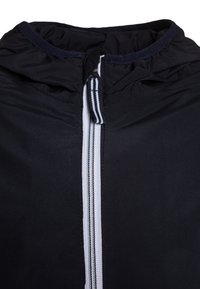 Chaqueta negra con cuello alto y cremallera, con acentos de rayas blancas a lo largo de la cremallera. Tejido suave con una textura ligeramente brillante.