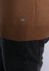 Brun strikket sweater med ribbet kant, prydet med et metal logo mærke. Kombineret med sorte denim bukser, der viser tekstureret stof.
