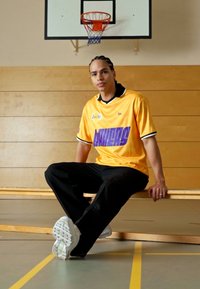 Gelbes Basketball-Trikot mit lila Aufschrift, schwarzem Rand; lockere schwarze Hose; weiße Turnschuhe; sitzt an einer holzverkleideten Wand der Turnhalle.