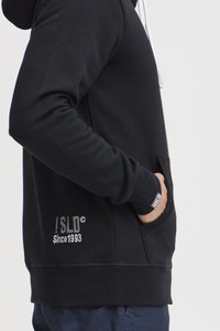 Sudadera negra con bolsillo frontal, cordones blancos, puños acanalados y detalles de logo bordado en el cuerpo y las mangas.