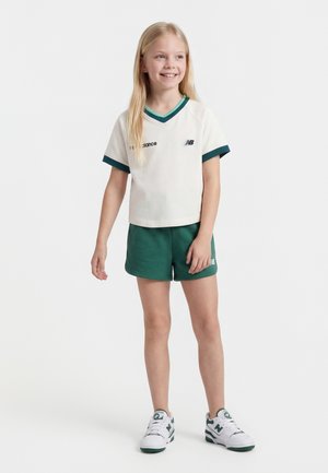 Ung pige står og smiler, iført en hvid kortærmet skjorte med grøn kant, grønne shorts og hvide sneakers med grønne detaljer.