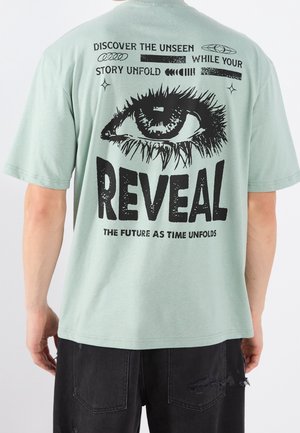 Mint-vihreä puuvillapaita, jossa on musta graafinen printti silmästä ja teksti takana. Paidassa on lyhyet hihat ja rento istuvuus.