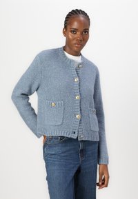 Lichtblauwe gebreide cardigan met een ronde halslijn, gouden knopen, geribde boorden en twee voorzakken, gedragen met spijkerbroek.