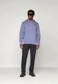 Vīrietis, kurš valkā purpura kapuci, melnus bikses, melnus sporta apavus un lielas melnas saules brilles, stāv pretī vienkāršai gaišai fonam.