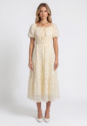 Frau mit blondem Haar, die ein hellgelbes Midi-Kleid mit Puffärmeln, eine geraffte Taille, weiße Absätze und goldenen Schmuck trägt, stehend vor weißem Hintergrund.
