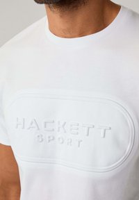Bílý bavlněný tričko s vyvýšeným logem 'HACKETT SPORT' uprostřed. Kulatý výstřih a krátké rukávy, hladká textura.